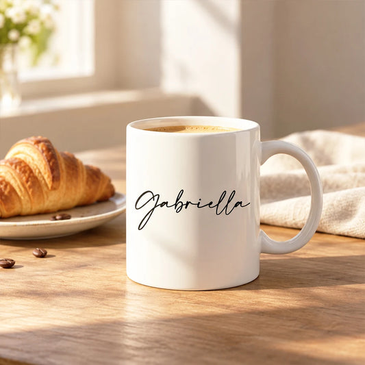 Tazza bianca personalizzata con il nome Gabriella accanto a una colazione con cornetto